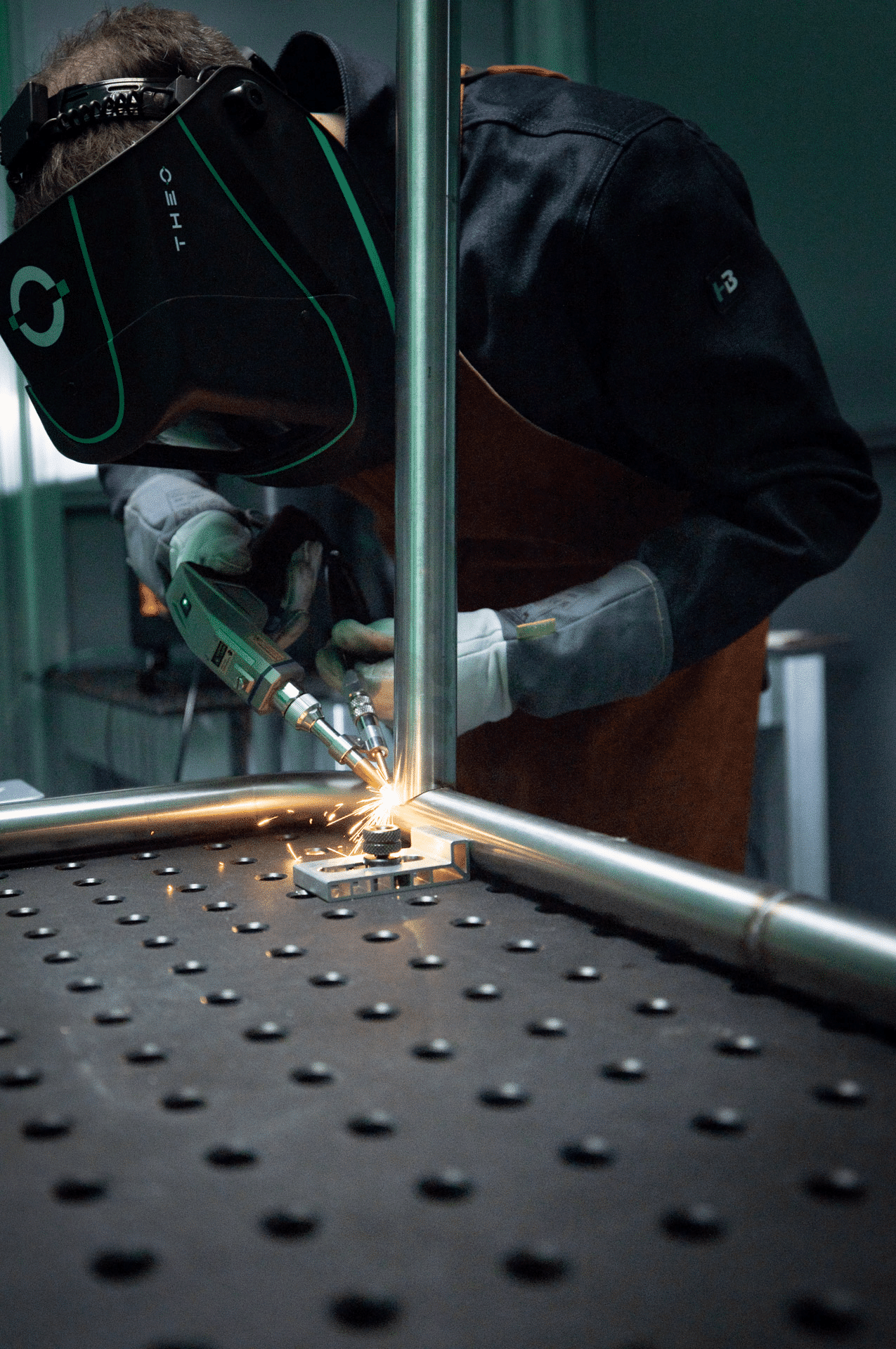 Iförd Laserklassad svetshjälm THEO VIZ-R2 svetsar en person metallrör med ett elverktyg på en perforerad arbetsbänk, med kraftiga gnistregn vid kontaktpunkten.