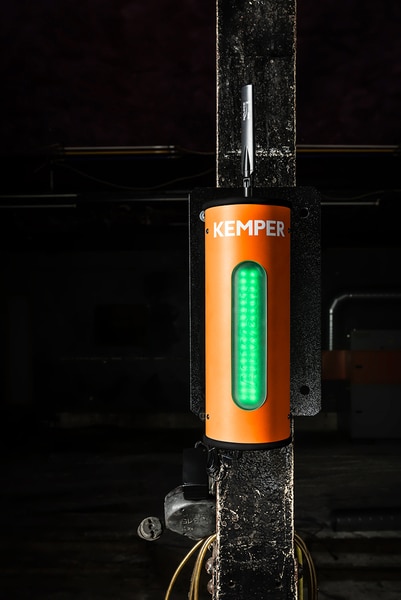 En Kemper luftkvalitetsövervakning AirWatch, med ett vertikalt grönt ljus på frontpanelen, är monterad vertikalt på en mörk stolpe med synliga kablar och rör nedanför. Bakgrunden verkar svagt upplyst.