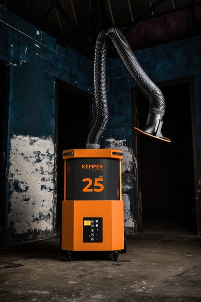 En orange industrimaskin märkt "Kemper mobilt filterutsug SmartFil" står på ett betonggolv, med en flexibel utsugningsslang som sträcker sig uppåt mot en mörk, sliten blå och vit vägg.