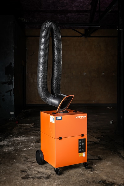 Kemper mobilt rökutsug SmartMaster är en industriorange luftfiltreringsenhet på hjul med en flexibel svart utblåsningsslang som sträcker sig uppåt, i ett svagt upplyst, oavslutat rum med betonggolv och betongväggar.